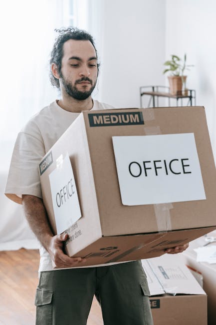 A person wearing a yellow t-shirt and blue jeans is standing inside a room with light-colored walls and wooden flooring, surrounded by cardboard boxes, some open and empty, ready for packing. The indi