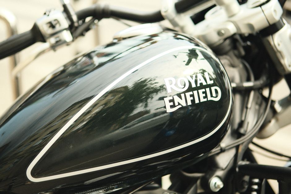 A black and red Royal Enfield motorcycle parked on a paved road in front of a lush green field, with a backdrop of dense trees and a cloudy sky. The motorcycle features a classic design with a red fue