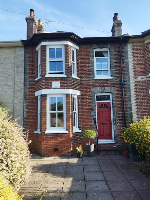 A traditional two-storey red brick Victorian terrace house with white window frames and a red front door, situated in Enfield Town on a sunny day. In the foreground, there is a concrete paved pathway