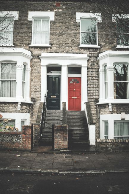 A traditional two-storey red brick Victorian terrace house with white window frames and a red front door, situated in Enfield Town on a sunny day. In the foreground, there is a concrete paved pathway