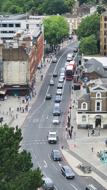 An aerial view of a busy urban street with parked and moving cars, including vans and small vehicles, alongside a row of retail and residential buildings. The street has a designated lane for vehicles